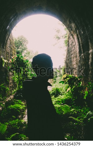 Picture of a silhouette of a man standing inside of an old abandoned metro tunnel which being overgrowth with plants and sun light at the end