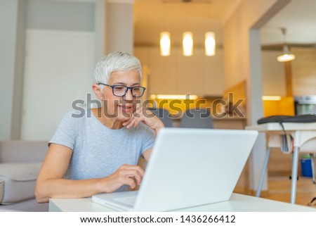 Elderly woman working on laptop computer, smiling. Woman Working From Home On Laptop In Modern Apartment. Trendy woman working on laptop from home