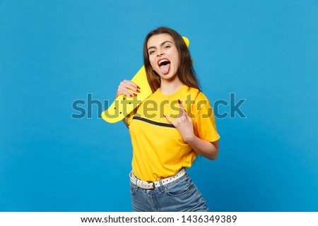 Portrait of crazy funny young woman in vivid casual clothes hold yellow skateboard showing tongue, horns up gesture isolated on blue background in studio. People lifestyle concept. Mock up copy space