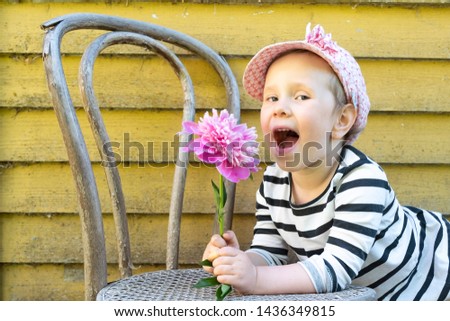 Portrait of a child, a girl in a cap with a peony flower in her hands. child on the background of old yellow woman, wooden wall with an old vienna chair.
