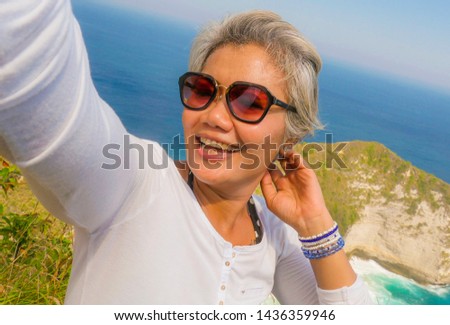 middle age 50s happy and cheerful Asian woman with grey hair taking selfie with mobile phone at beautiful tropical beach island smiling at cliff viewpoint enjoying Summer holidays travel destination
