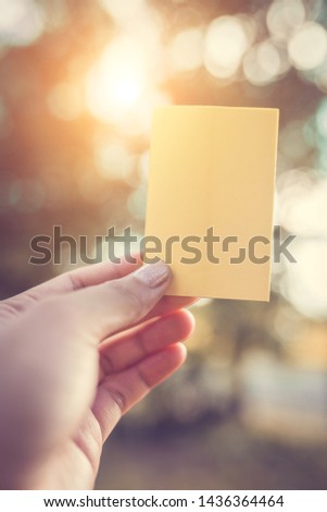 The hand is holding a color paper with empty space for note with a backdrop of sunlight and natural bokeh.