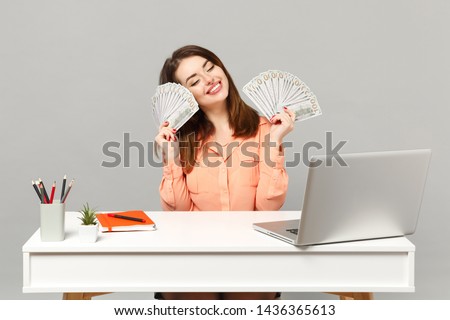 Young charming woman holding fan of cash money in dollar banknotes, sit and work at desk with pc laptop isolated on gray background. Achievement business career lifestyle concept. Mock up copy space