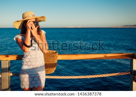 Young woman traveler taking photos of sea landscape on pier using camera. Summer fashion