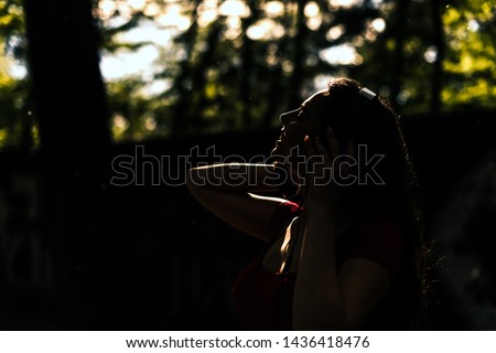 Silhouette of fit girl listening to music streaming with headphones in summer in the park, low key, dark image