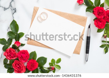 Wedding invitation card, craft paper envelope, bridal rings, pen, rose flowers arrangement on marble desk table. Flat lay style composition, top view, overhead. Wedding invitation card mockup. 