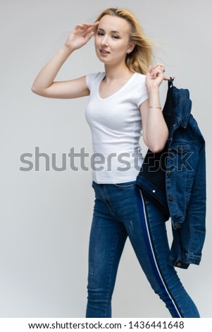 Portrait of pretty interesting fashionable blonde girl on a white background in jeans and a blue jacket. Standing in front of the camera, smiling, showing hands. Shows a lot of emotions.