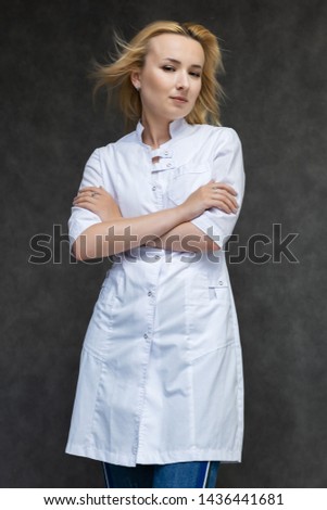 Portrait of a knee-high pretty interesting beautiful girl blonde doctor on a gray background in a white medical coat. Standing in front of the camera, smiling, a lot of emotions.