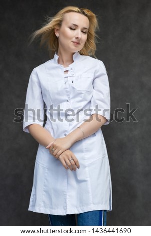 Portrait of a knee-high pretty interesting beautiful girl blonde doctor on a gray background in a white medical coat. Standing in front of the camera, smiling, a lot of emotions.