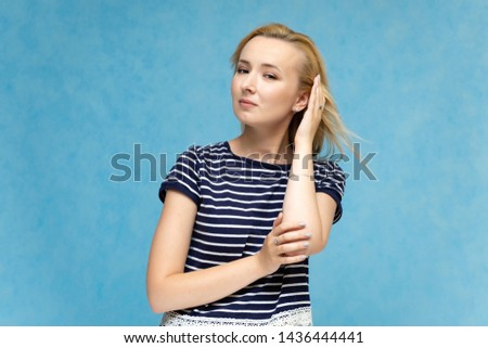 Portrait to the waist of a nice interesting fashionable blonde girl on a blue background in a striped blue t-shirt. Standing in front of the camera, smiling, showing hands. Shows a lot of emotions.