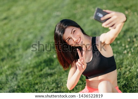 young beautiful sport brunette girl in sports clothes sit on the grass after a workout and making selfie on a smart phone. Healthy lifestyle concept.