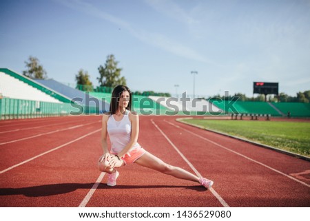 Beautiful fitness brunette girl in sportswear and sneakers does a stretching exercise on the running track at the stadium outdoors. Sun shines onto her body.