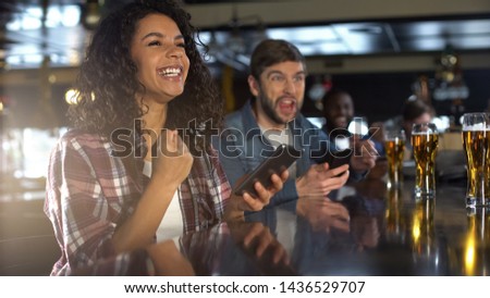 Smiling biracial woman in pub celebrating winning sports bet, bookmaker app Royalty-Free Stock Photo #1436529707