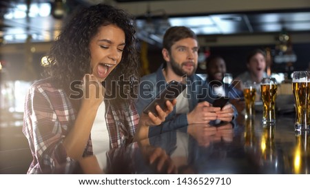 Cheerful biracial lady in pub celebrating successful bet on sports, online app Royalty-Free Stock Photo #1436529710
