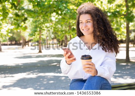 Happy woman drinking coffee and using smartphone on bench. Beautiful lady wearing blouse and sitting in park with green trees in background. Communication and rest concept. Front view.