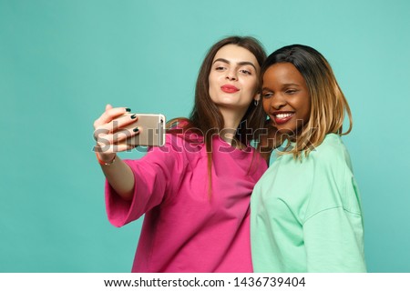 Two women friends european and african american in pink green clothes doing selfie shot on cellphone isolated on blue wall background, studio portrait. People lifestyle concept. Mock up copy space