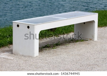 New smart bench in local public park used for charging mobile phones while providing wireless network and lightning gravel path next to uncut grass and lake on warm sunny spring day Royalty-Free Stock Photo #1436744945