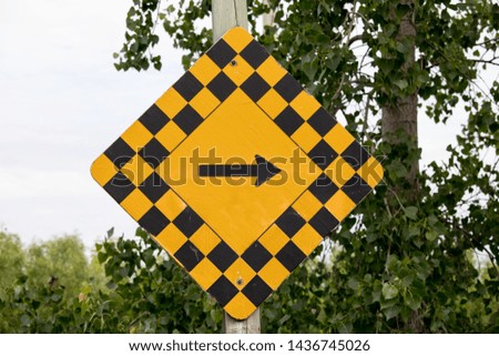 yellow and black checkerboard diamond road caution sign with black arrow pointing right near a tree