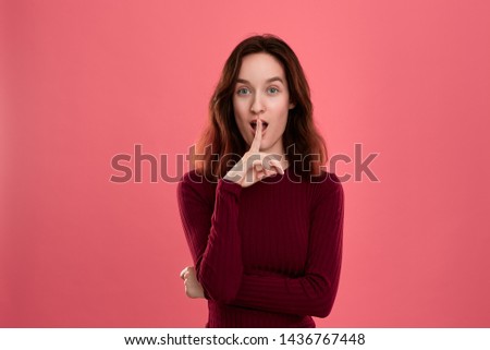 Beautiful young lady standing isolated over dark pink background with finger raised close to mouth expressing gesture of keeping silence and looking at the camera. Concept of secret