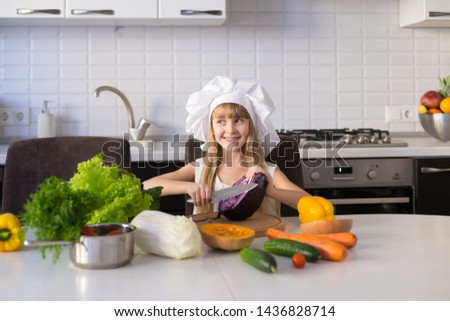 Beautiful little girl, has happy fun smiling face, long blonde hair, white chef hat. Cooks in kitchen appetizing tasty vegetables. Child portrait. Creative detox concept. Cuisine food view.