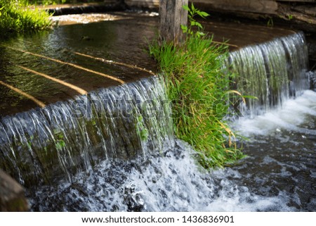 An artificial waterfall shaded by an aged overgrown wooden bridge on a sunny day.