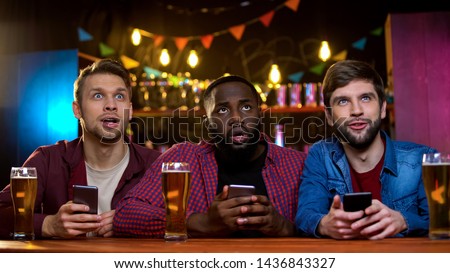 Mixed-race friends comparing lottery numbers on tv in bar with online tickets Royalty-Free Stock Photo #1436843327