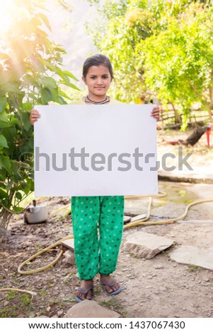 
Happy smiling little Indian village girl standing against in front a nature background. showing blank placard, whiteboard to write it on your own text