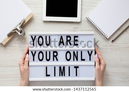 Female hands holding lightbox with 'You are your only limit' words over white wooden background, top view. 