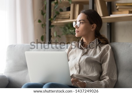 Dreamy millennial woman sit on couch hold laptop look in distance thinking distracted from online work, thoughtful girl in glasses take break from computer studying dreaming or visualizing at home Royalty-Free Stock Photo #1437249026