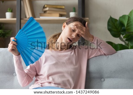 Overheated millennial woman sit on couch at home feel warm waving with hand fan cooling down, sweating girl relax on sofa in living room hold waver suffer from heat, no air conditioner system Royalty-Free Stock Photo #1437249086