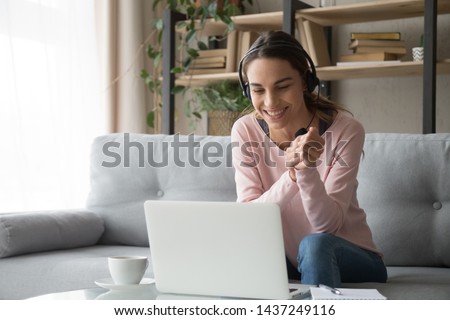 Smiling millennial girl in headset sit on couch watching webinar on laptop drinking coffee, happy young woman in earphones use computer on sofa in living room, study on online distant course Royalty-Free Stock Photo #1437249116