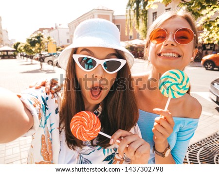 Two young smiling hipster women in casual summer clothes. Girls taking selfie self portrait photos on smartphone.Models posing on street background in sunglasses and panama hat.Eating candy lollipop