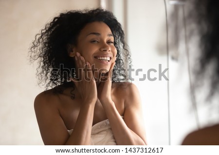 Close up black woman after shower stands wrapped in towel look in mirror touch gentle skin feels happy after cleansing mask, prevention protection, antiaging treatment usage and natural beauty concept Royalty-Free Stock Photo #1437312617
