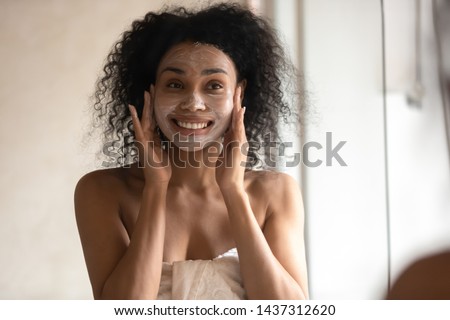 African 30s young woman after shower is wrapped in towel looking in mirror pampering herself applying face mask use anti-aging beauty product feeling happy, skin care, everyday morning routine concept Royalty-Free Stock Photo #1437312620