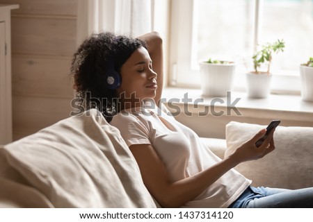 Attractive 30s mixed race woman wearing headphones closed her eyes holding smart phone listen enjoy favourite music lying in living room resting on couch spend lazy weekend at home, mood hobby concept Royalty-Free Stock Photo #1437314120