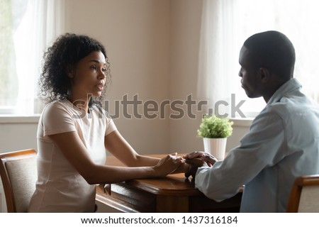 Serious african 30s married couple in love sitting at table having heart-to-heart intimate straight talk, mixed race wife holding hands of beloved black husband family share problems thoughts at home Royalty-Free Stock Photo #1437316184