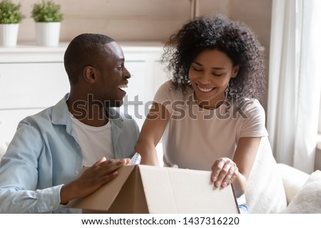 Satisfied couple relocates prepare belongings packing to carton big box moving day to new home. Black spouses opening received cardboard parcel package, easy and fast service commerce delivery concept Royalty-Free Stock Photo #1437316220