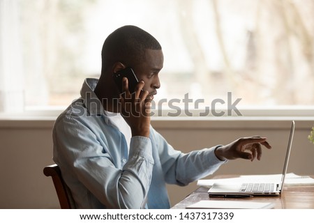 African 30s man sitting at table looking pointing at computer screen discuss application talking on mobile phone working at home solve business issues distantly, self-employed male speak with client Royalty-Free Stock Photo #1437317339