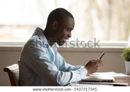 Side view black young man sit at table at home holds smart phone using having fun with apps, surfing internet browsing web sites, chatting distantly with friend, flirting use e-dating services concept
