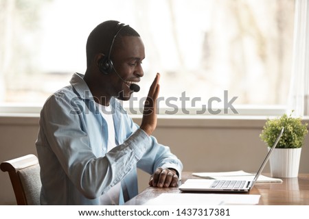 Black guy wear headset start lesson online look at laptop screen wave hand greeting tutor improves foreign language knowledge get skills through internet, education distantly using modern tech concept Royalty-Free Stock Photo #1437317381