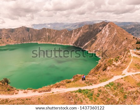 Dramatic DRONE shot perspective of Quilotoa lake and volcano crater, with view of mountains, hiking path trail loop and cloudy sky from viewpoint. Shot in Ecuador. Green and blue.