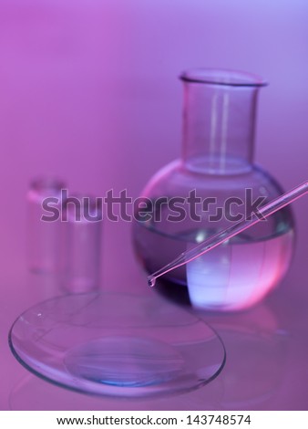 pipette dropping a transparent liquid in a dish with two empty beakers and a round boiling flask with transparent liquid in the background against a gradient purple background