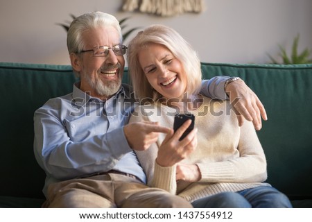 Happy senior old couple holding smartphone looking at cellphone screen laughing relaxing sit on sofa together, smiling elder mature grandparents family embracing having fun with mobile phone at home Royalty-Free Stock Photo #1437554912