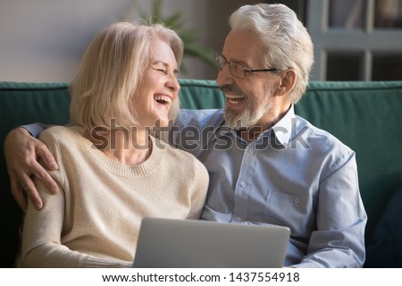Happy older mature spouses couple laughing embracing using laptop at home, positive cheerful senior middle aged family husband and wife bonding having fun with computer sitting on couch together
