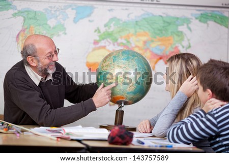 Senior male teacher pointing at globe while students looking at it at desk in classroom Royalty-Free Stock Photo #143757589