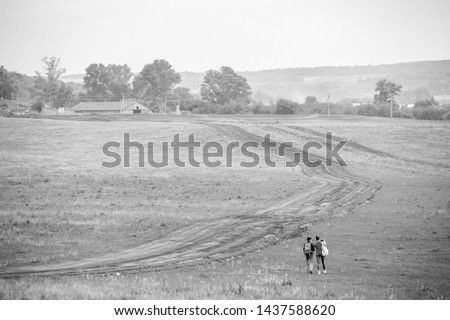 Two young girls with backpacks and photo camera make selfie on phone near rural road. Horse farm pasture with mare and foal. Small village with old houses. Summer landscape .Black and white