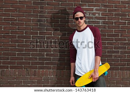 Urban fashion skateboarder with woolen hat and sunglasses in front of brick wall.