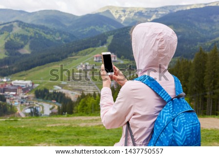 Back view of slender woman holding her smartphone in both hands, taking photos for travel blog, looking for new route online, searching for connection in mountain tour. Copyspace for advertisement.