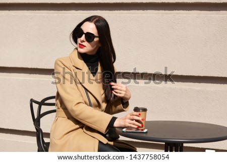 Horizontal shot of young beautiful brunette woman in stylish sunglasses and beige coat sitting at table in outdoor cafe, drinking hot baverage from paper cup, looking aside, touches her hair.