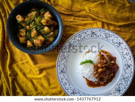 Top view flat lay image of a curry vegetable poured on rice in a white plate with a bowl of dry Indian style cooked potato on the side in a black bowl. 